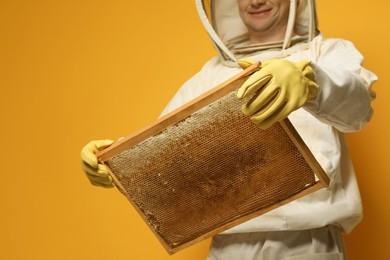 Beekeeper in uniform holding hive frame with honeycomb on yellow background, closeup