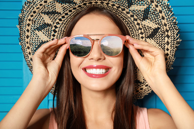 Young woman wearing stylish sunglasses with reflection of mountains and hat on blue wooden background 