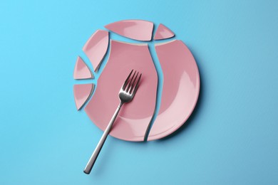 Pieces of broken ceramic plate and fork on light blue background, flat lay