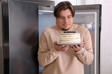 Happy overweight man with cake near open refrigerator in kitchen