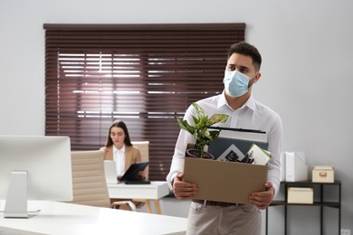 Upset dismissed man wearing protective mask carrying box with personal stuff in office