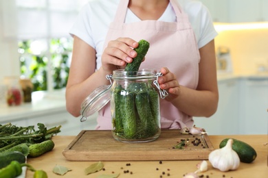 Woman putting cucumber into pickling jar at table in kitchen, closeup