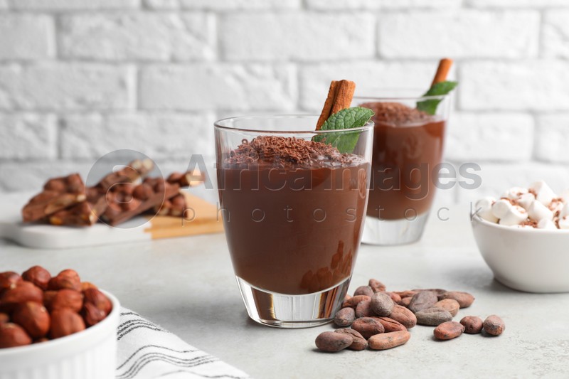 Photo of Cups of delicious hot chocolate with cinnamon stick, mint and cocoa beans on light grey table near brick wall