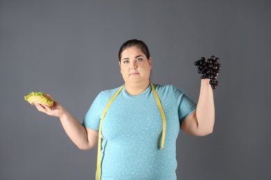 Overweight woman with hamburger, grapes and measuring tape on gray background
