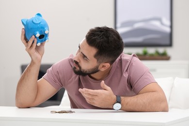 Worried young man with piggy bank and money at white table indoors