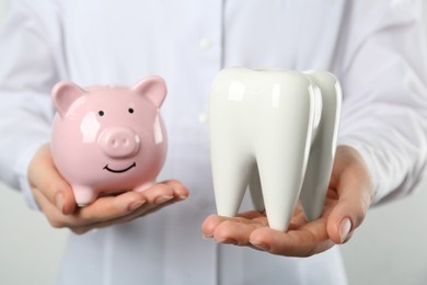 Dentist holding ceramic model of tooth and piggy bank on light background, closeup. Expensive treatment