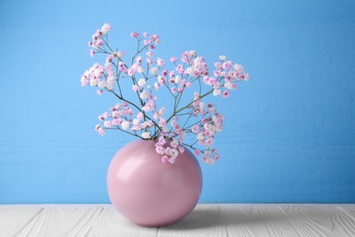 Photo of Beautiful dyed gypsophila flowers in pink vase on white wooden table against light blue background