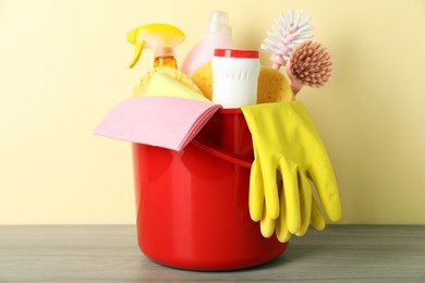 Bucket with different cleaning supplies on wooden floor near beige wall, closeup
