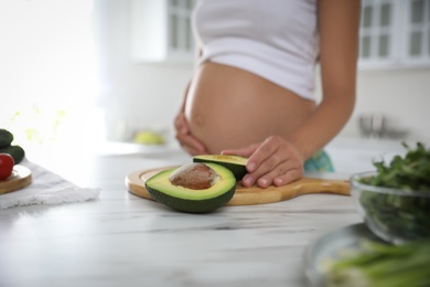 Young pregnant woman with avocado at table in kitchen, closeup. Taking care of baby health