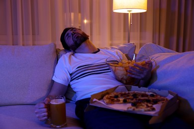 Photo of Man with chips, pizza and glass of beer sleeping on sofa at night. Bad habit