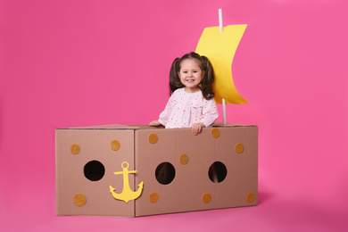 Photo of Little child playing with ship made of cardboard box on pink background