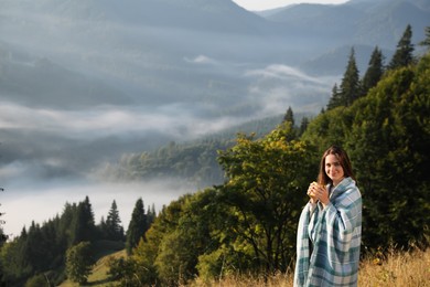Young woman with cup of hot drink and plaid enjoying beautiful mountain landscape. Space for text