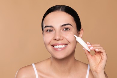 Beautiful young woman applying gel on skin under eye against beige background