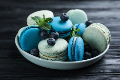 Delicious macarons, mint and blueberries in bowl on black wooden table, closeup