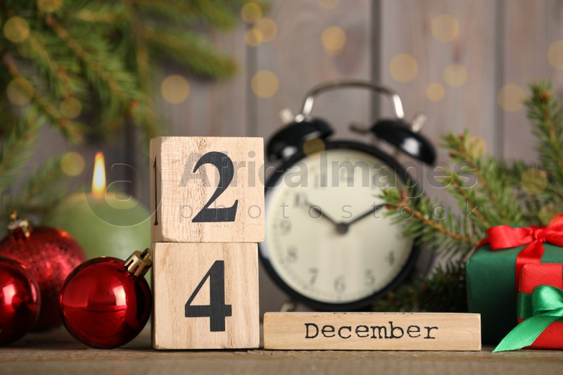 Photo of December 24 - Christmas Eve. Wooden block calendar, alarm clock and festive decor on table against blurred lights