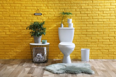 Stylish bathroom with toilet bowl, green plants and decor elements near yellow brick wall. Interior design