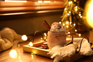 Tasty hot drink with whipped cream and Christmas lights on wooden table