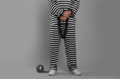 Prisoner in special uniform with chained hands and metal ball on grey background, closeup