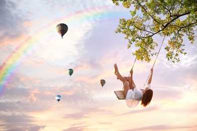 Dream world. Young woman swinging, hot air balloons in sunset sky on background