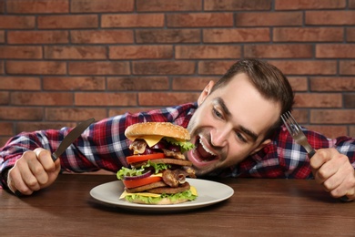 Young hungry man with cutlery eating huge burger at table