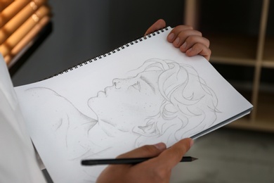 Man drawing portrait with pencil in notepad indoors, closeup