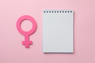 Photo of Female gender sign as women's health symbol and notebook on pink background, flat lay. Space for text