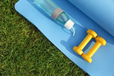 Photo of Dumbbells, bottle of water and fitness mat on green grass, flat lay. Morning exercise