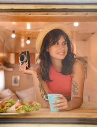 Young woman with cup and vintage camera in trailer, view from outside. Camping vacation