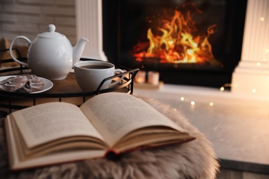 Cup of tea, cookies and book on faux fur near fireplace indoors, space for text. Cozy atmosphere