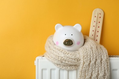 Piggy bank wrapped in scarf and thermometer on heating radiator against orange background