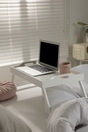 White tray table with laptop and cup of drink on bed indoors