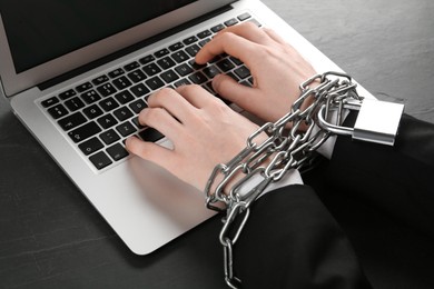 Photo of Internet addiction. Man typing on laptop with chained hands at black table, closeup