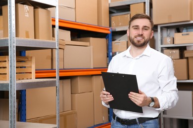 Young businessman with clipboard near rack of cardboard boxes at warehouse