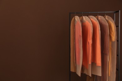 Photo of Garment bags with clothes on rack against brown background. Space for text