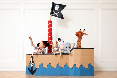 Cute little boy playing with toys in pirate cardboard ship near white wall indoors