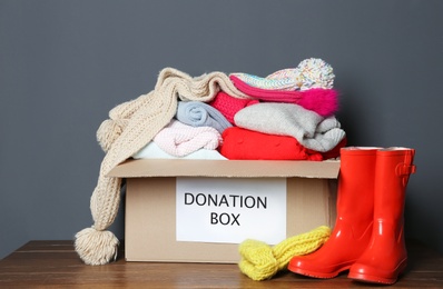 Donation box with knitted clothes and rubber boots on table near grey wall