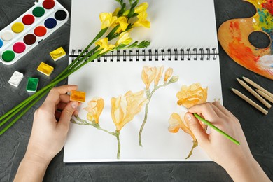 Woman drawing beautiful freesia flowers in sketchbook at black table, above view
