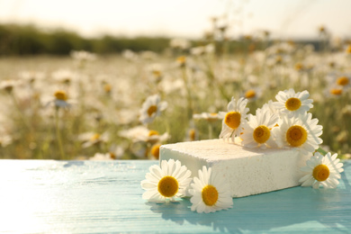 White soap bar and fresh chamomiles on light blue wooden table in field. Space for text