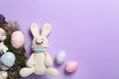 Toy bunny with protective mask, painted eggs and space for text on lilac background, flat lay. Easter holiday during COVID-19 quarantine