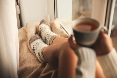 Woman in knitted socks holding cup of tea on plaid near window at home, closeup