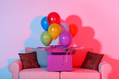 Gift box with bright air balloons on sofa against color background
