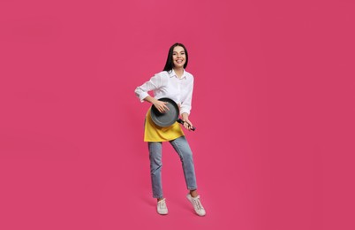 Young housewife with frying pan having fun on pink background