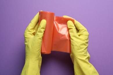 Photo of Janitor in rubber gloves holding roll of orange garbage bags over purple background, top view