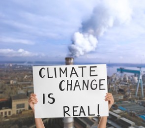 Protestor holding placard with text Climate Change Is Real and blurred view of industrial factory on background 