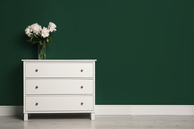 Photo of Modern white chest of drawers with beautiful bouquet near green wall indoors. Space for text