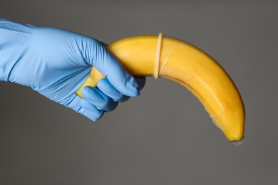 Doctor holding banana in condom symbolizing male sexual organ on grey background, closeup. Potency problem