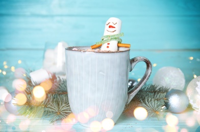 Funny marshmallow snowman in cup of hot drink on light blue wooden table. Bokeh effect