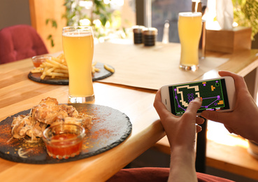Woman playing game using smartphone at table with tasty BBQ wings in cafe, closeup