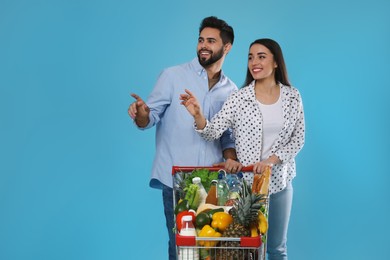 Young couple with shopping cart full of groceries on light blue background