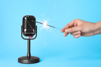 Woman making ASMR sounds with microphone and burning sparkler on light blue background, closeup